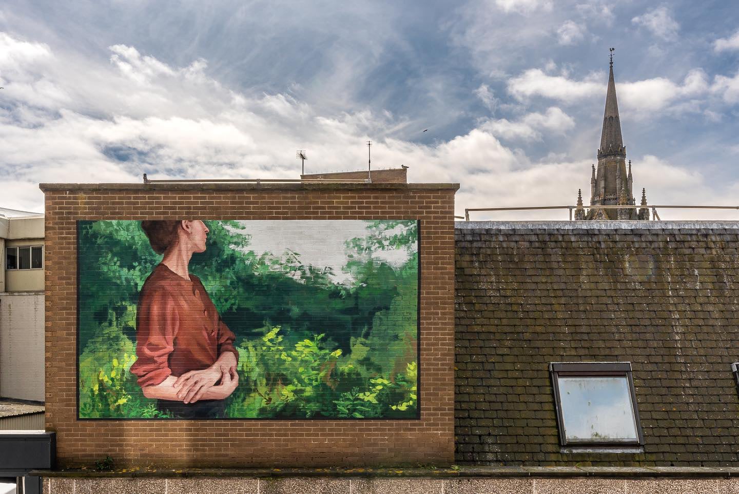 A woman wearing a beige top with her face turned away from the viewer looks back at lush greenery and clasps her arms in a mural on a brick facade.  A bell tower is in the background