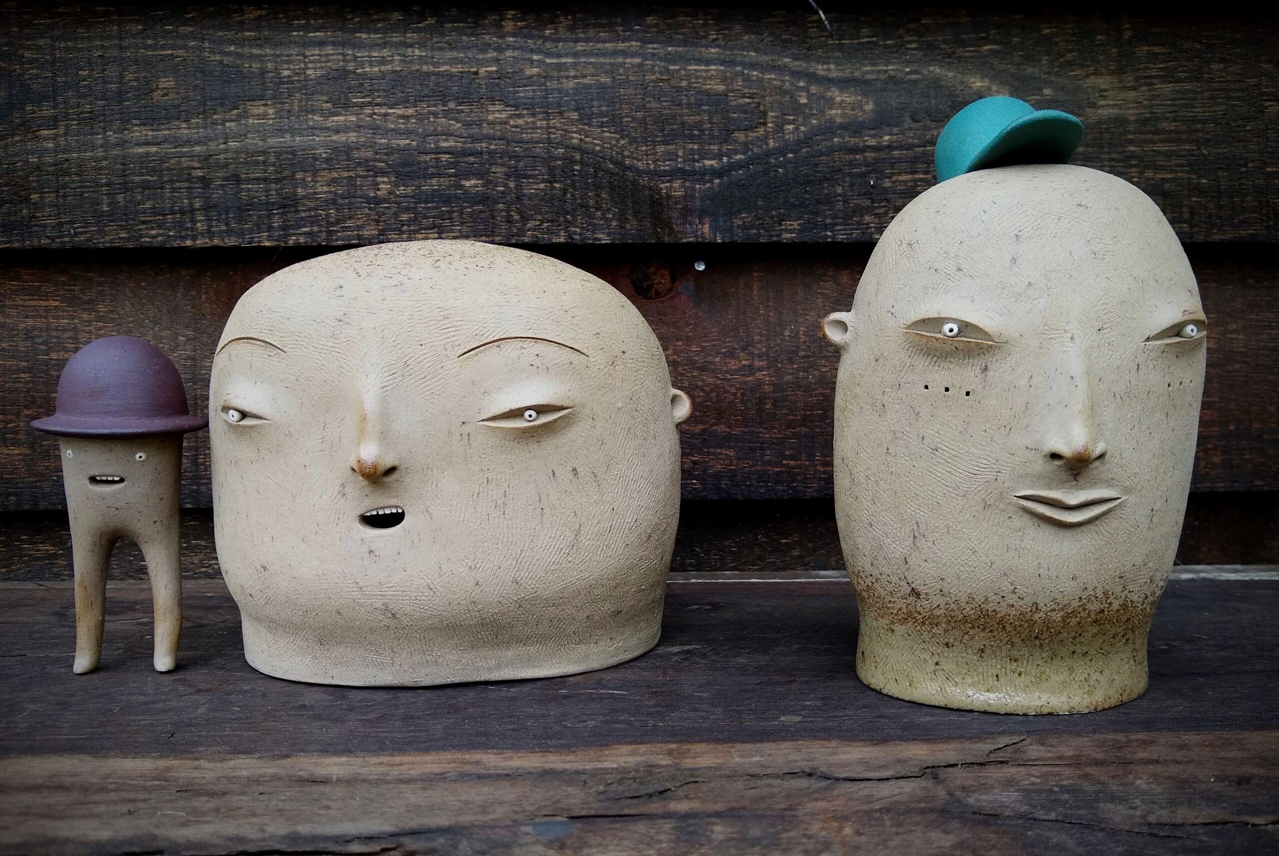 Two large ceramic heads sit next to each other on a wooden surface. A small figure with only two legs stands to the left, wearing a small hat. The large head on the right wears a small hat as well.