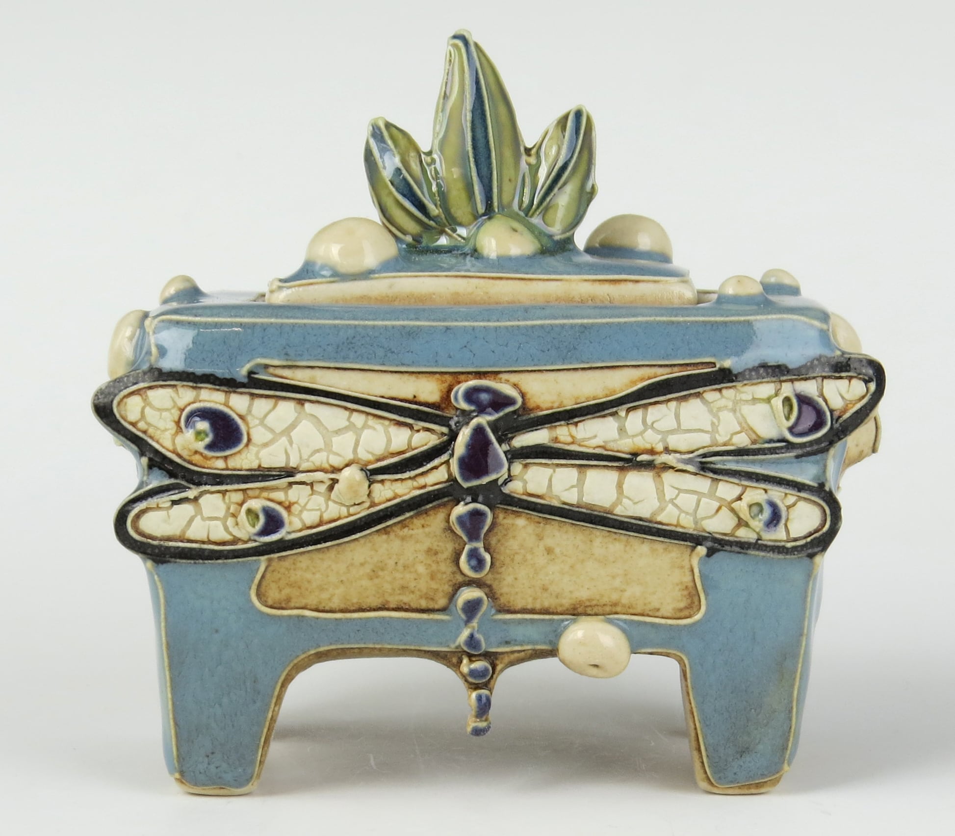 a dragonfly with mosaic wings adorns a ceramic box-like work with green leaves at the top