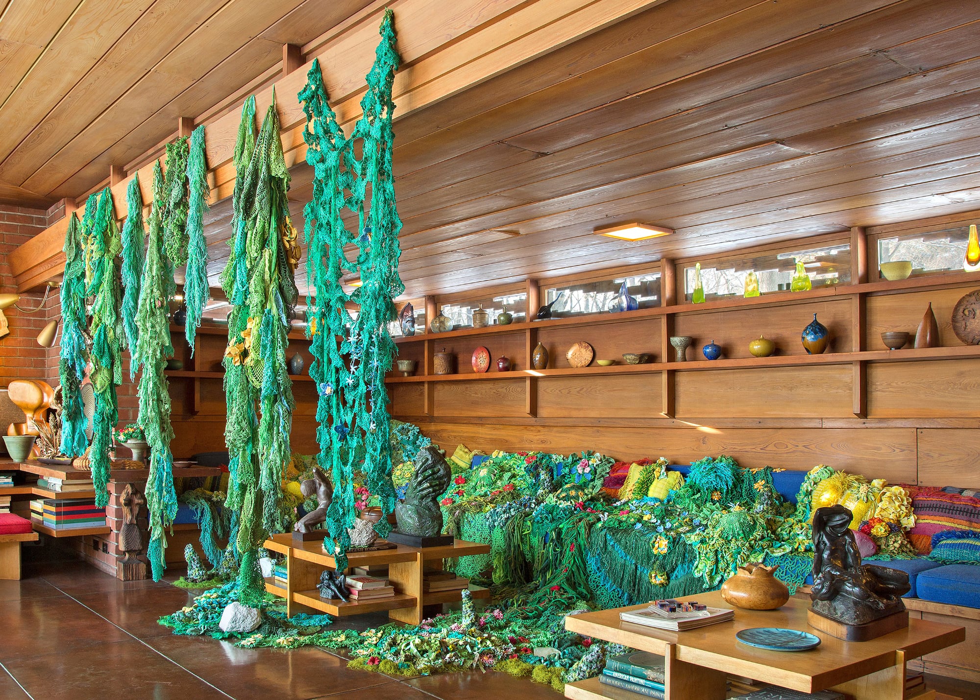 green crocheted forms hang from a wooden ceiling with crocheted flora on a nearby bench