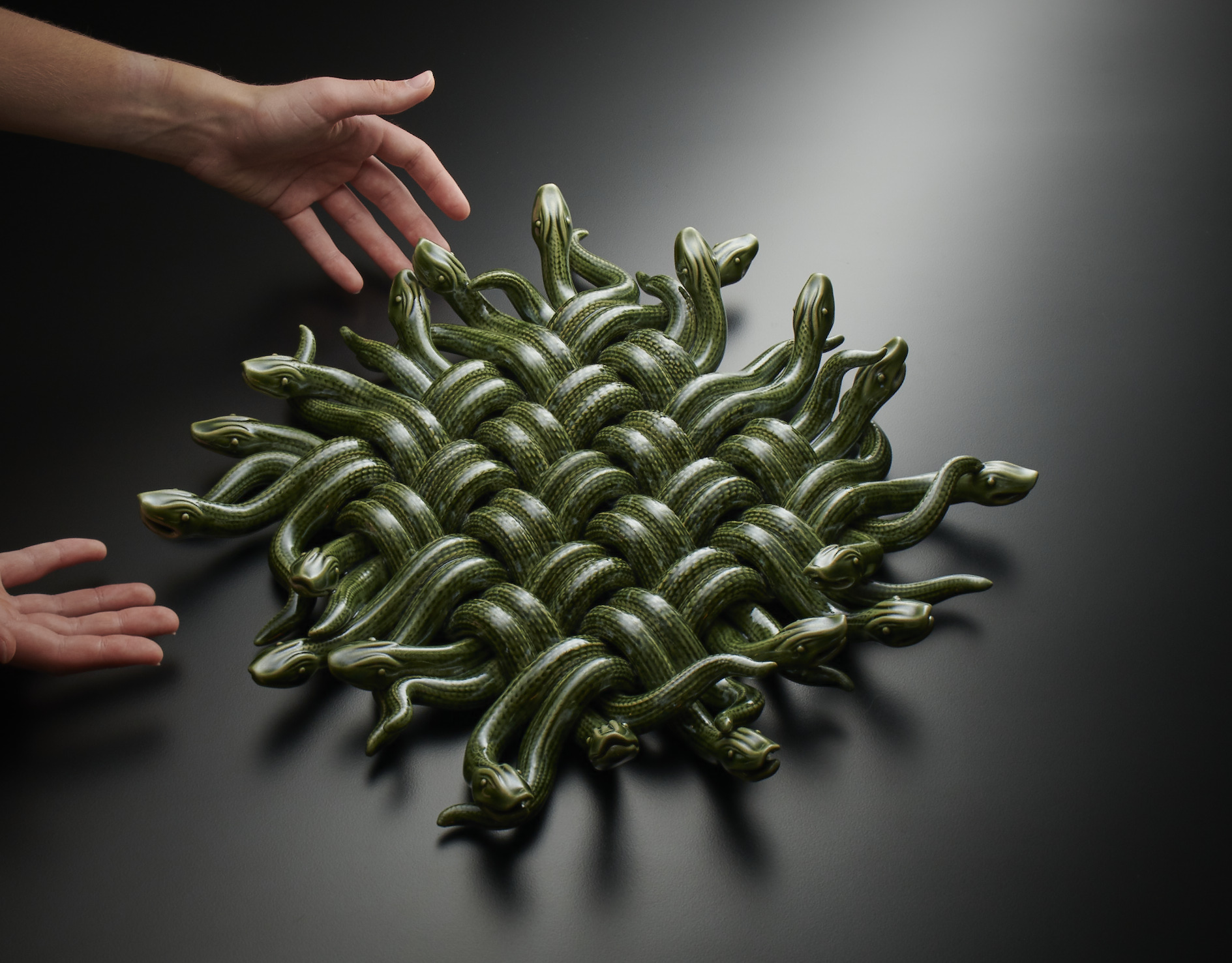 A ceramic sculpture of snakes woven into a mat. Photographed on a black background with hands reaching for it.