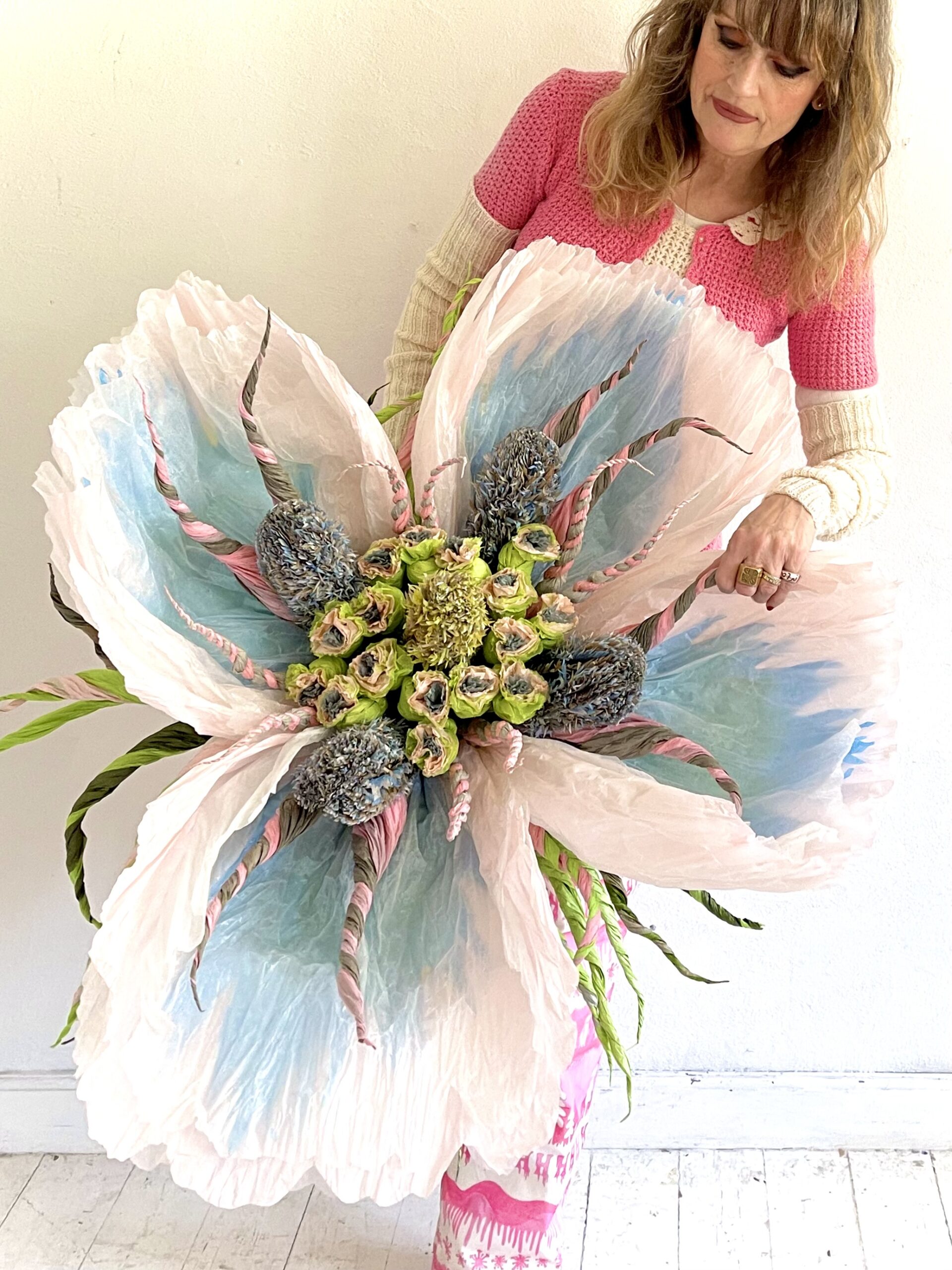 The artist is shown holding a large paper sculpture of a flower with green and pink interior and petals of blue and white.