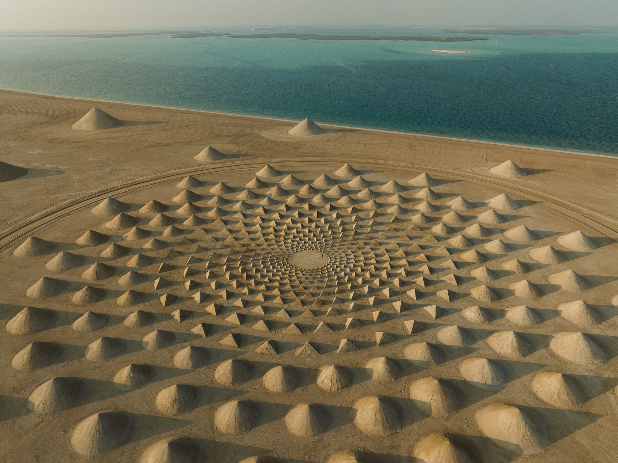 An aerial view of a giant land art installation featuring 19 concentric circles of 448 pyramids formed from sand. The installation is on the shore of the Arabian Sea, and a figure stands in the center. 