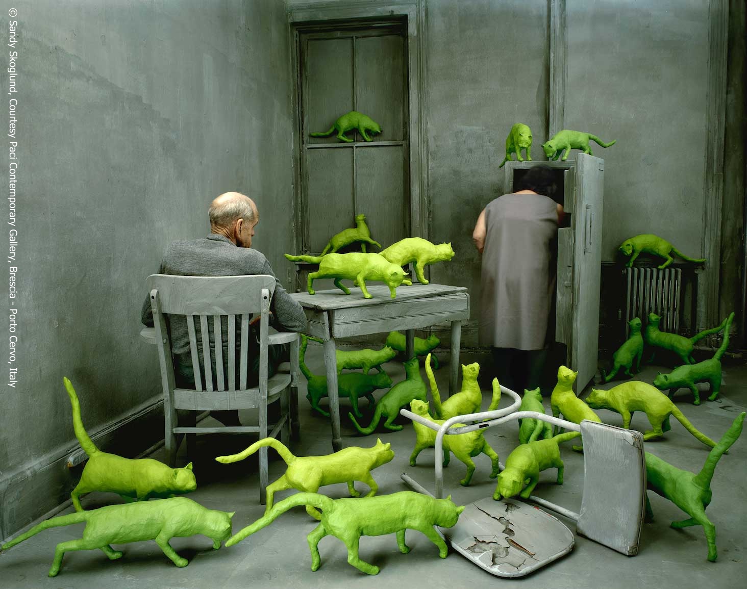 A fine art photograph of a gray kitchen interior with two elderly figures. A man is seated a table and a woman peers away from the viewer into a fridge. Numerous bright green cats run around.