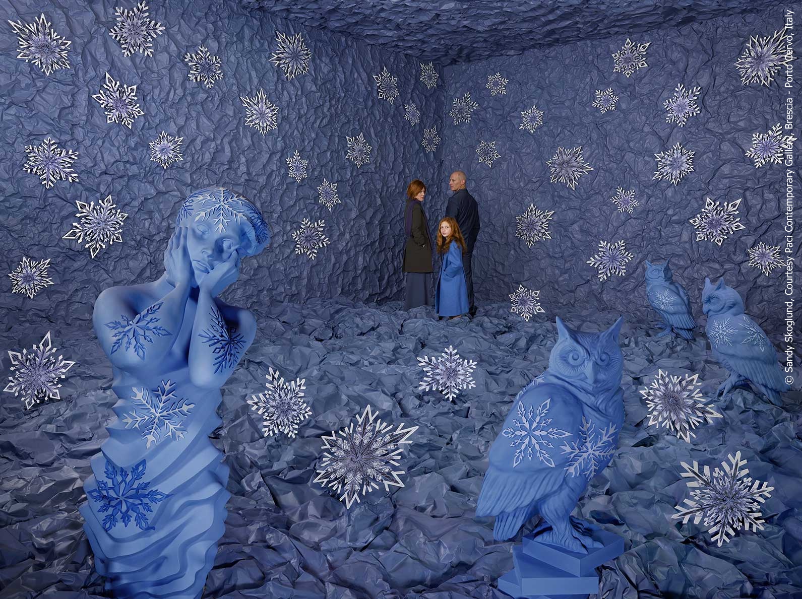 A surreal fine art photograph of a family in the corner of a room, coated in blue paper with snowflakes everywhere. In the foreground are figures of a young girl and some owls, also with snowflakes on them.