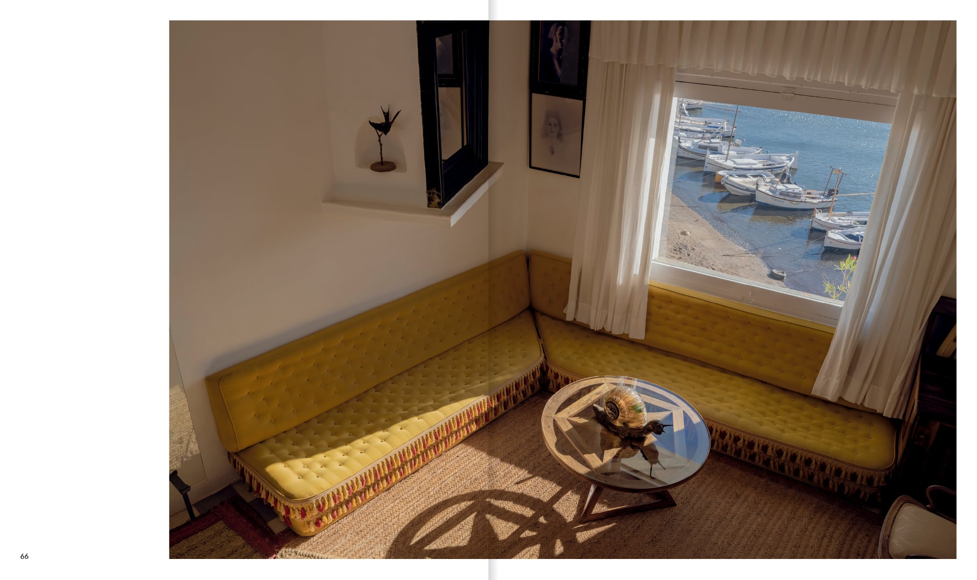 a book spread open to one image of a yellow L-shaped couch with a window on the right side looking down to the beach with small fishing boats docked