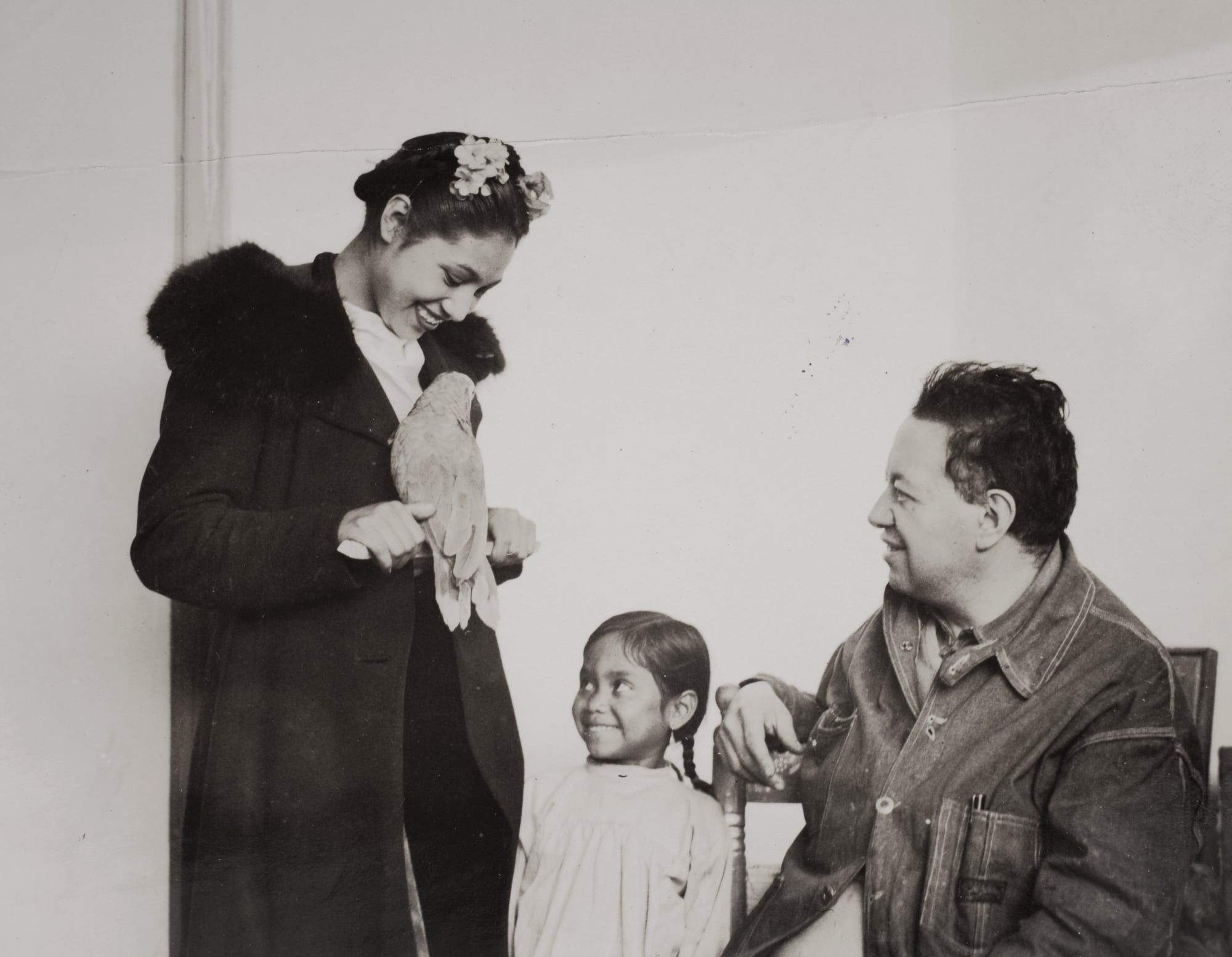 a woman wearing a fur-collared coat holds a bird while a young girl looks up at it with a smile. a man with short hair sits in a chair nearby