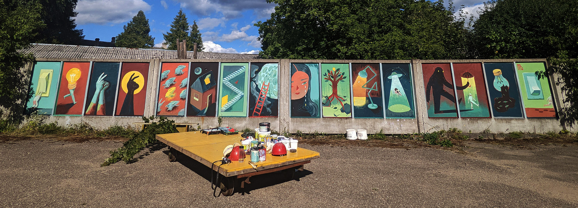 A wide shot of a mural depicting 16 panels, each with a different scene. In the foreground sits a cart with painting supplies.