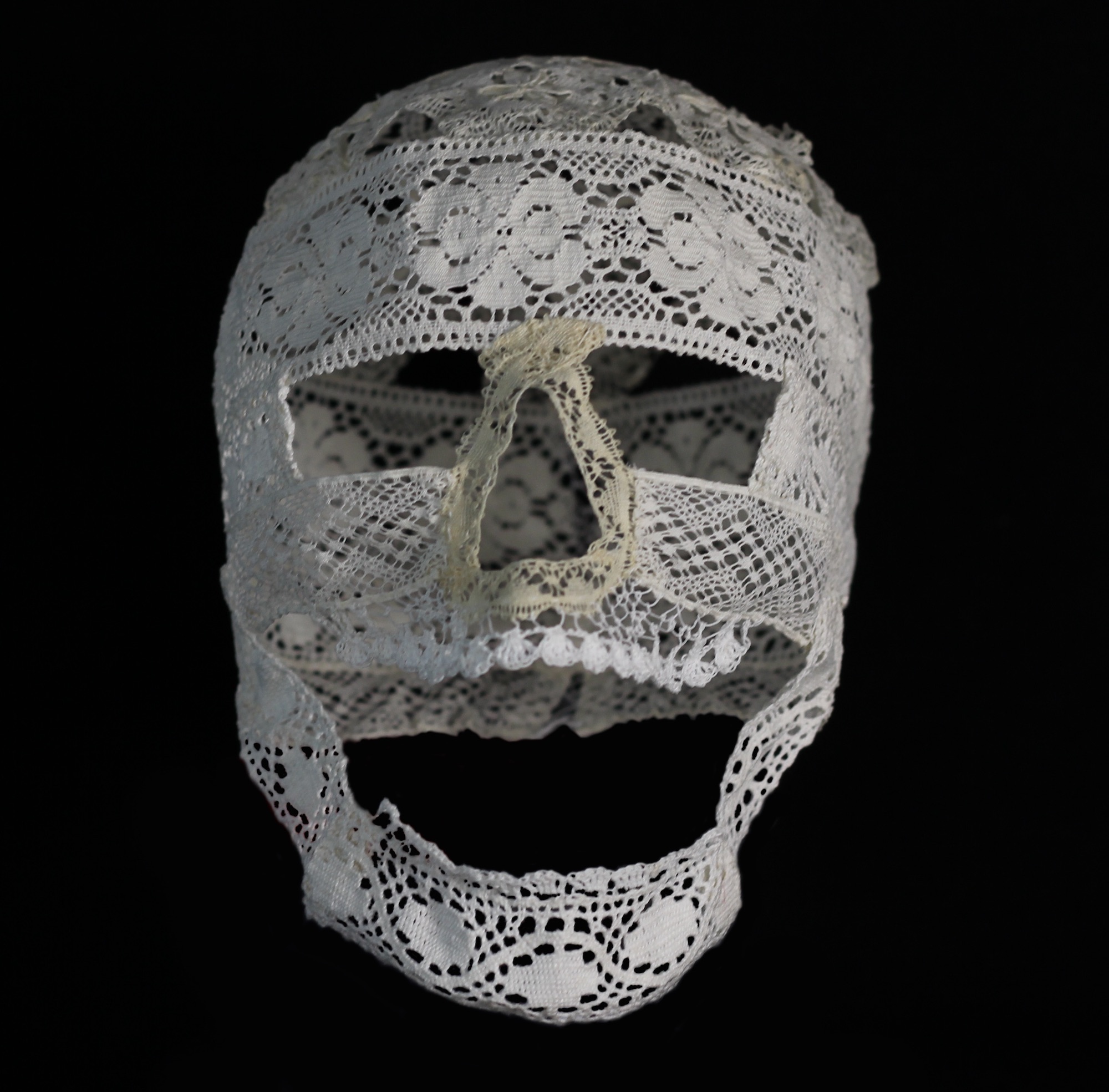 A sculpture of a human skull made entirely from piece of lace sewn together. It is photographed on a black background and shown facing forward.