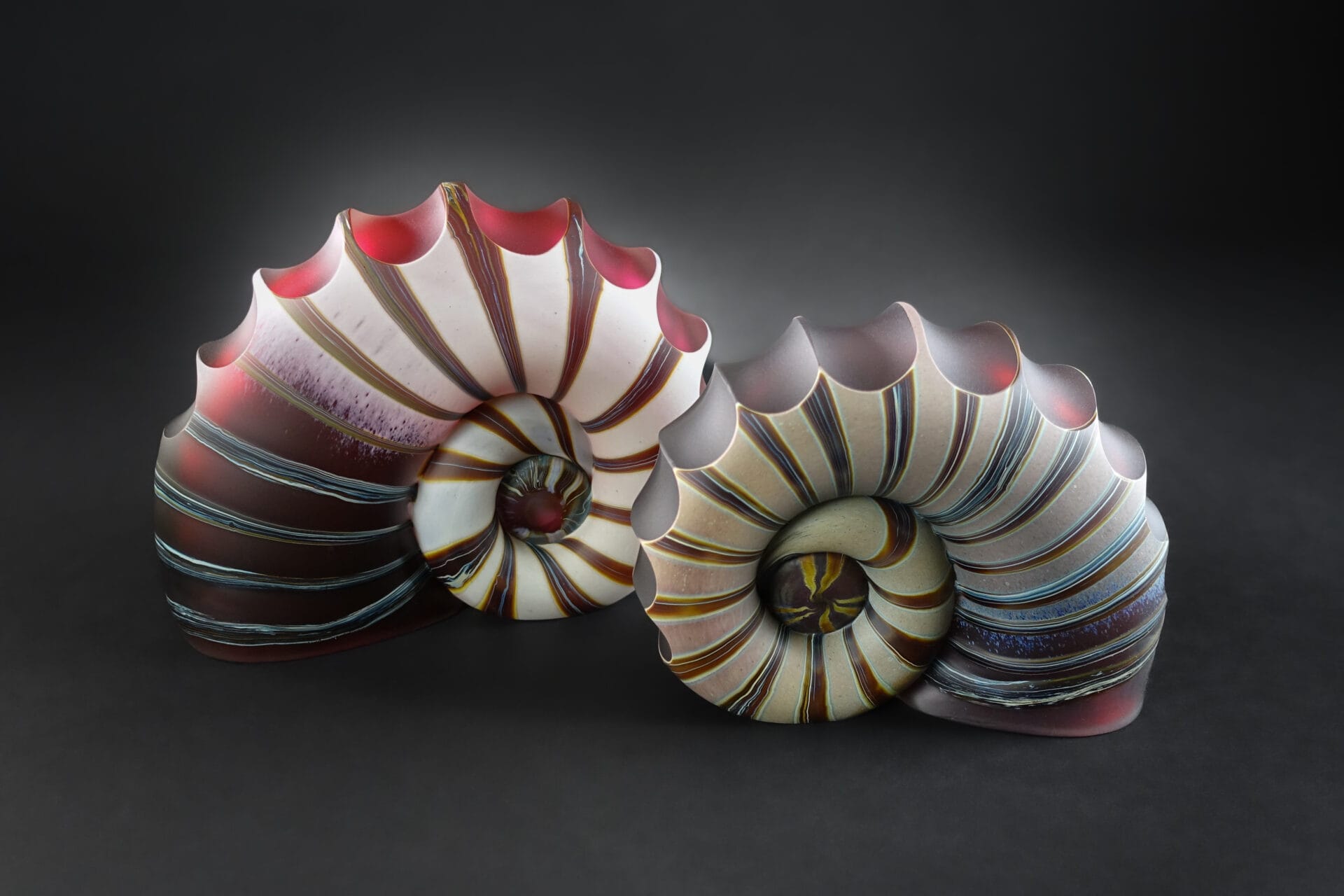 Two glass ammonites, in their unique spiral shell, in hues of ivory, red, and blue.